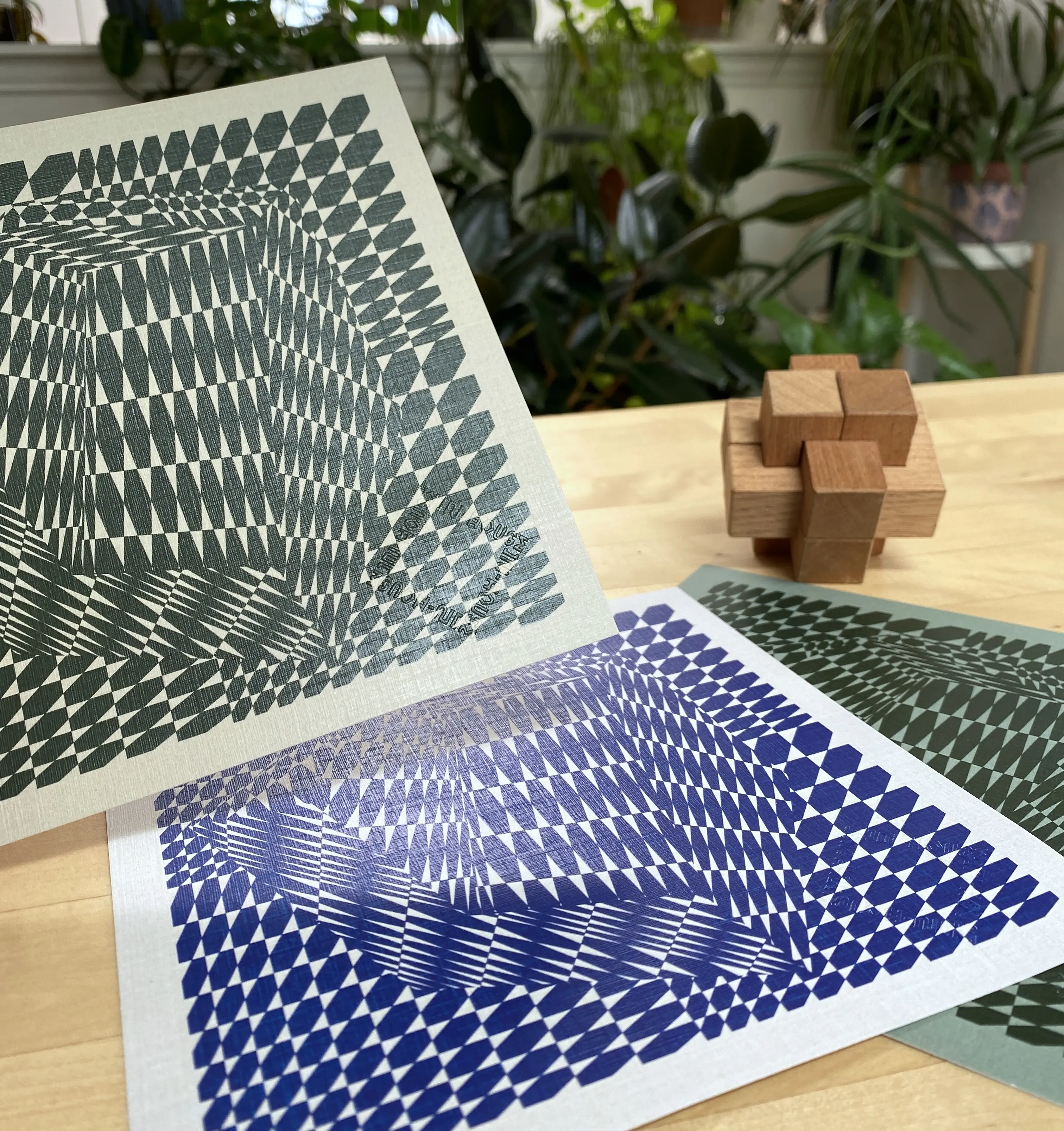 Photo of three graphic prints, two green and one blue, on square card stock, depicting isometric faceted designs by In-House International. The prints are displayed on a wooden tabletop. There's plants in the background.